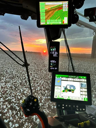 Interior de um trator, com o fundo da lavoura durante a colheita de algodão, com telas de controle mostrando dados agrícolas e um pôr do sol ao fundo