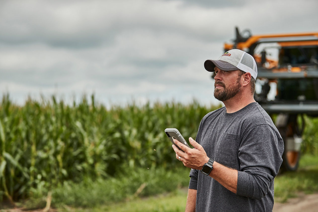 Farmer holding a smartphone, standing in a cornfield with farming equipment in the background under cloudy skies.