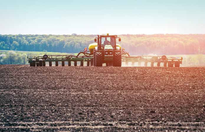 John Deere planter driving in the distance of a field of dark brown soil. Trees behind planter with blue sky above. 