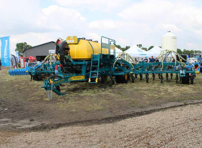 Blue and yellow Equalizer tine seeder with liquid bin on display on a dirt and grass patch at a tradeshow. A gravel road in the foreground with tradeshow tents, flags and a building in the background.