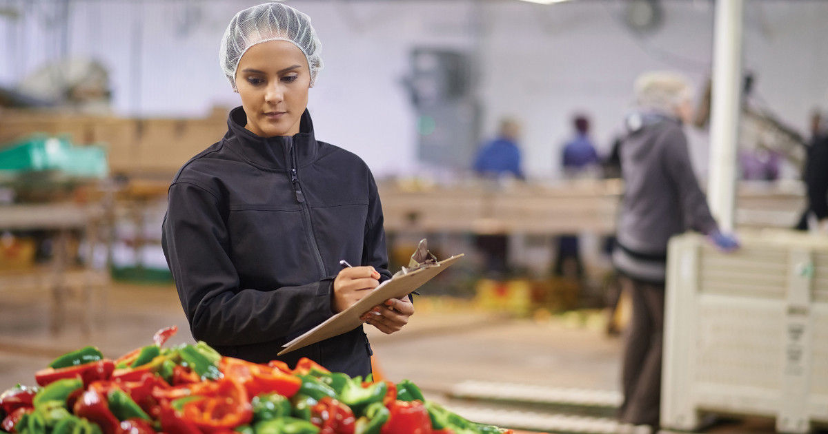 A food inspector holding a clipboard and pen, standing in front of a large pile of assorted red and green bell peppers. The background shows an indoor facility with other workers and equipment, indicating a food processing or inspection area.