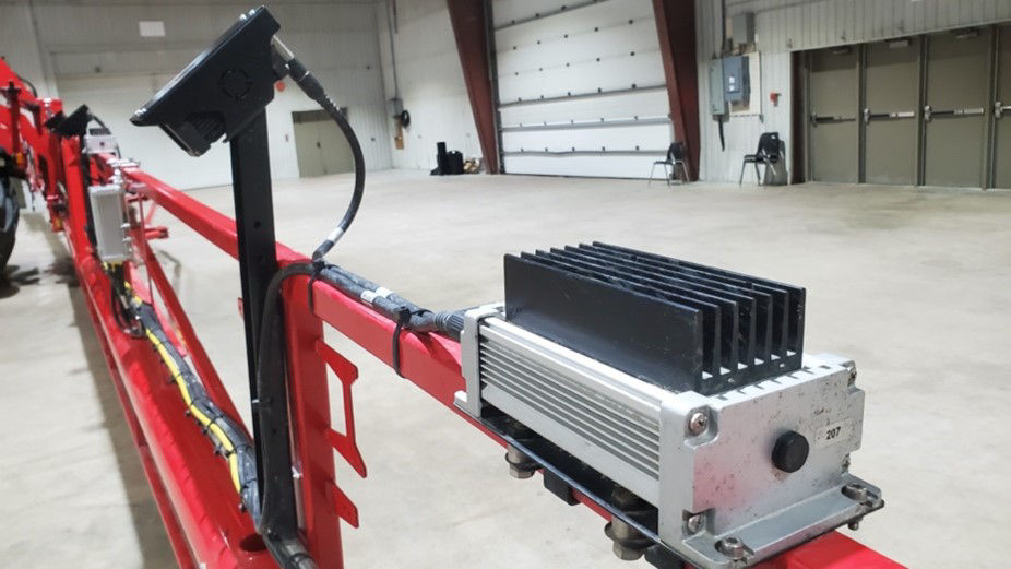 A black camera system mounted on the arm of a large red sprayer that’s being stored in a large empty building. 