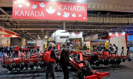 Two people stand in front of a large piece of agriculture machinery displayed at a tradeshow. There is a red sign that says “Canada”.