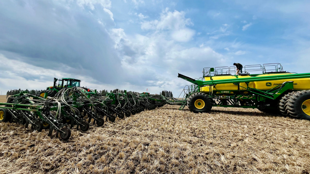 A wide-angle photograph of agricultural machinery, including a large John Deere air seeder and tractor, operating in a field under a partly cloudy sky. One person is seen on top of the seed tank, emphasizing the scale and complexity of the equipment.