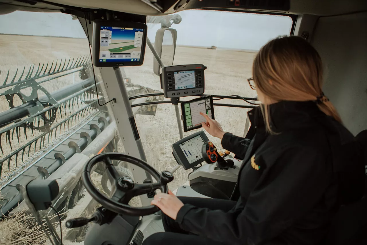 Woman operating a combine harvester, using several mounted monitors and touchscreens inside the cab while a header cuts across a dry field.