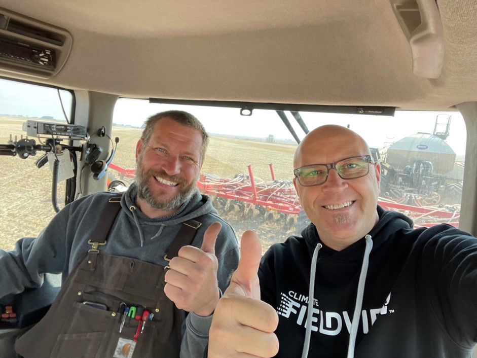 Two men sit in the cab of a tractor side by side, both smiling and giving the thumbs up sign. 