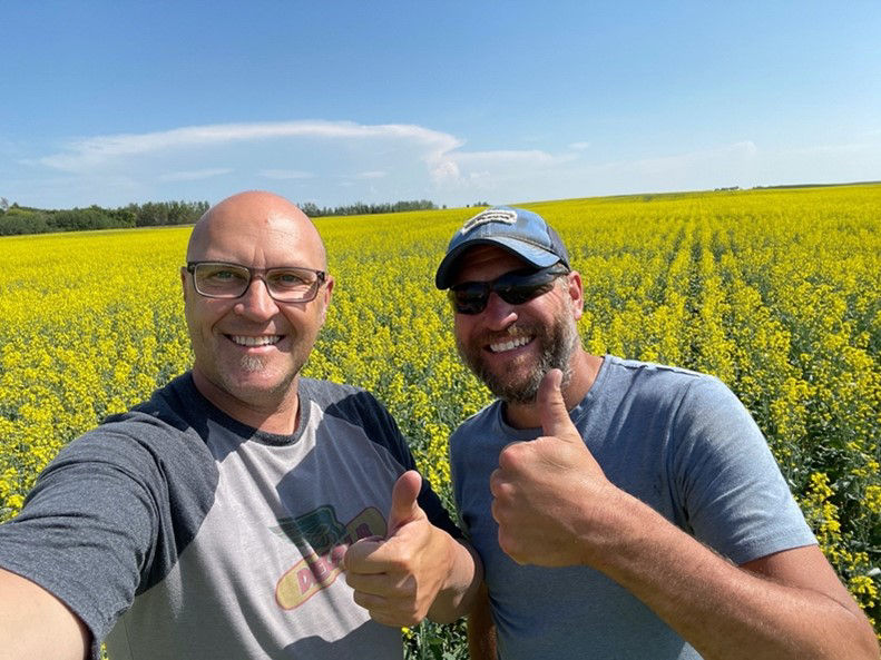 Two men stand in front of a thriving bright yellow canola field. They are both smiling and giving the thumbs up sign.