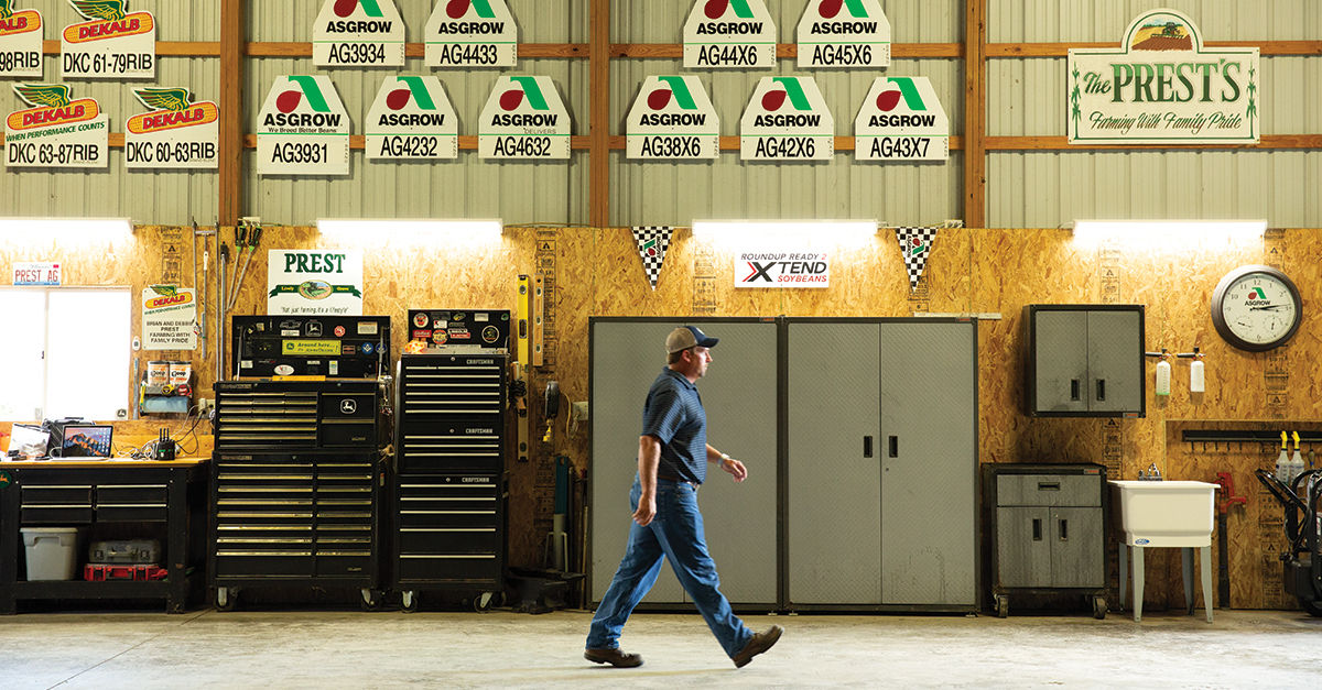 A farmer walking in a warehouse with Asgrow, Dekalb, Roundup Ready Xtend logos and The Prest's Family Farming signs hanging on the