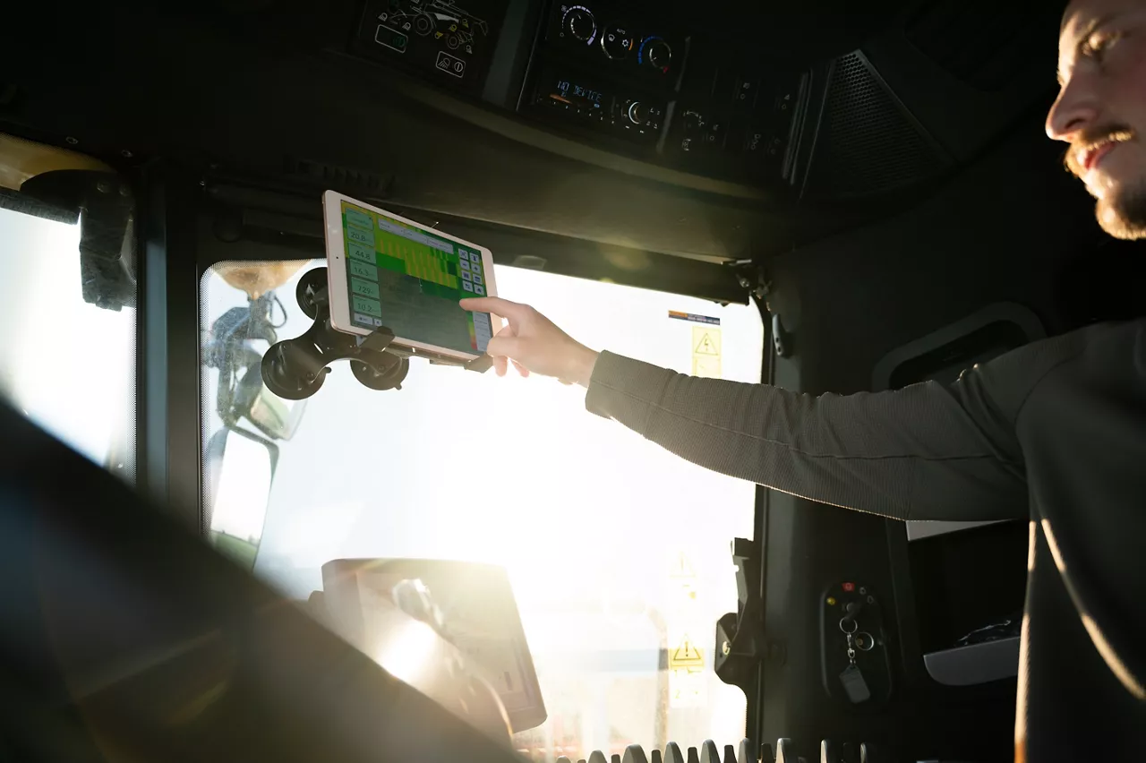 Farmer sitting in a tractor cab reaching forward to tap a tablet mounted on the windshield, with bright sunlight streaming through the window.