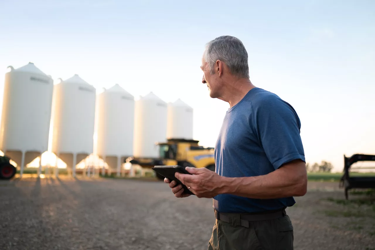 Un agriculteur âgé debout dans une cour de ferme au coucher du soleil tient une tablette et observe une rangée de silos à grains blancs et une moissonneuse-batteuse garée à l'arrière-plan.