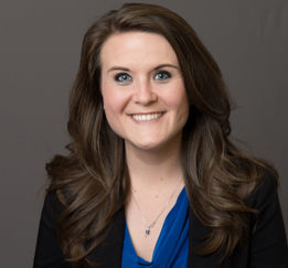 Professional headshot of a woman with long brown hair wearing a black blazer over a blue top against a grey background