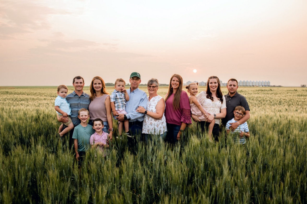 A large Caucasian family of seven adults and six children stand in a sprawling field of wheat, with silos along the distant horizon.