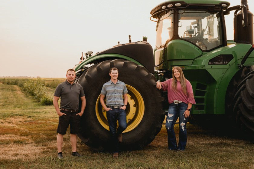 Two men and one woman stand leaning against the wheel of a giant green tractor parked at the edge of a sprawling wheat field. 