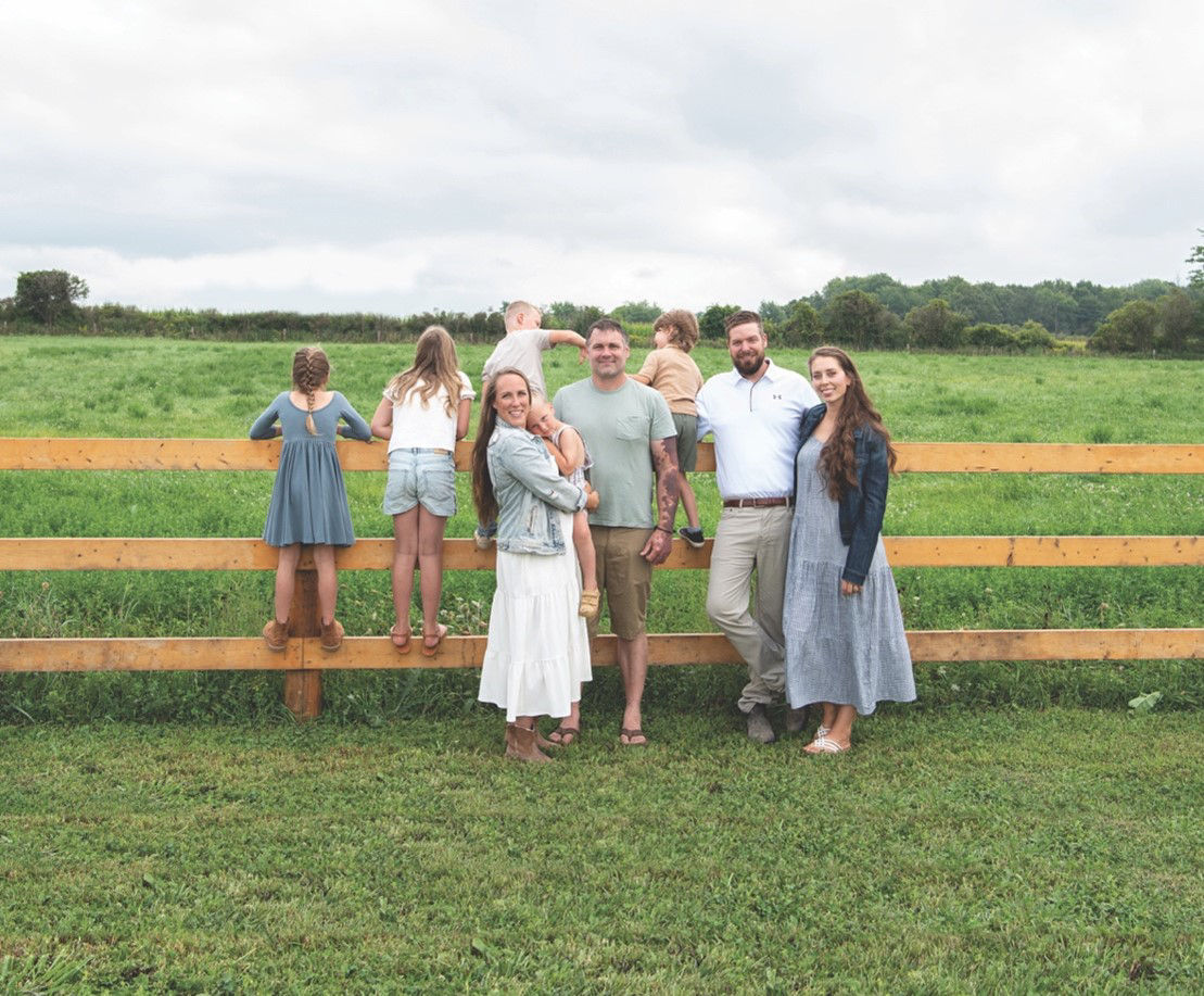 Two couples pose for a photo in front of a wooden fence. Several children climb on the fence behind them. 