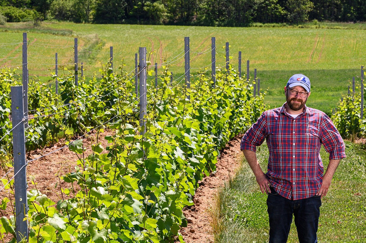 Mike Mainguy stands in a vineyard, posing with hands on hips for a photo. He is wearing a red flannel shirt and a casual baseball hat.