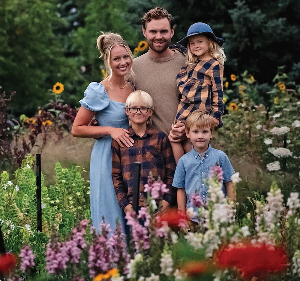 A family of five stands close together in a lush garden surrounded by colorful flowers. The parents stand behind their three young children, and they are all smiling at the camera.  