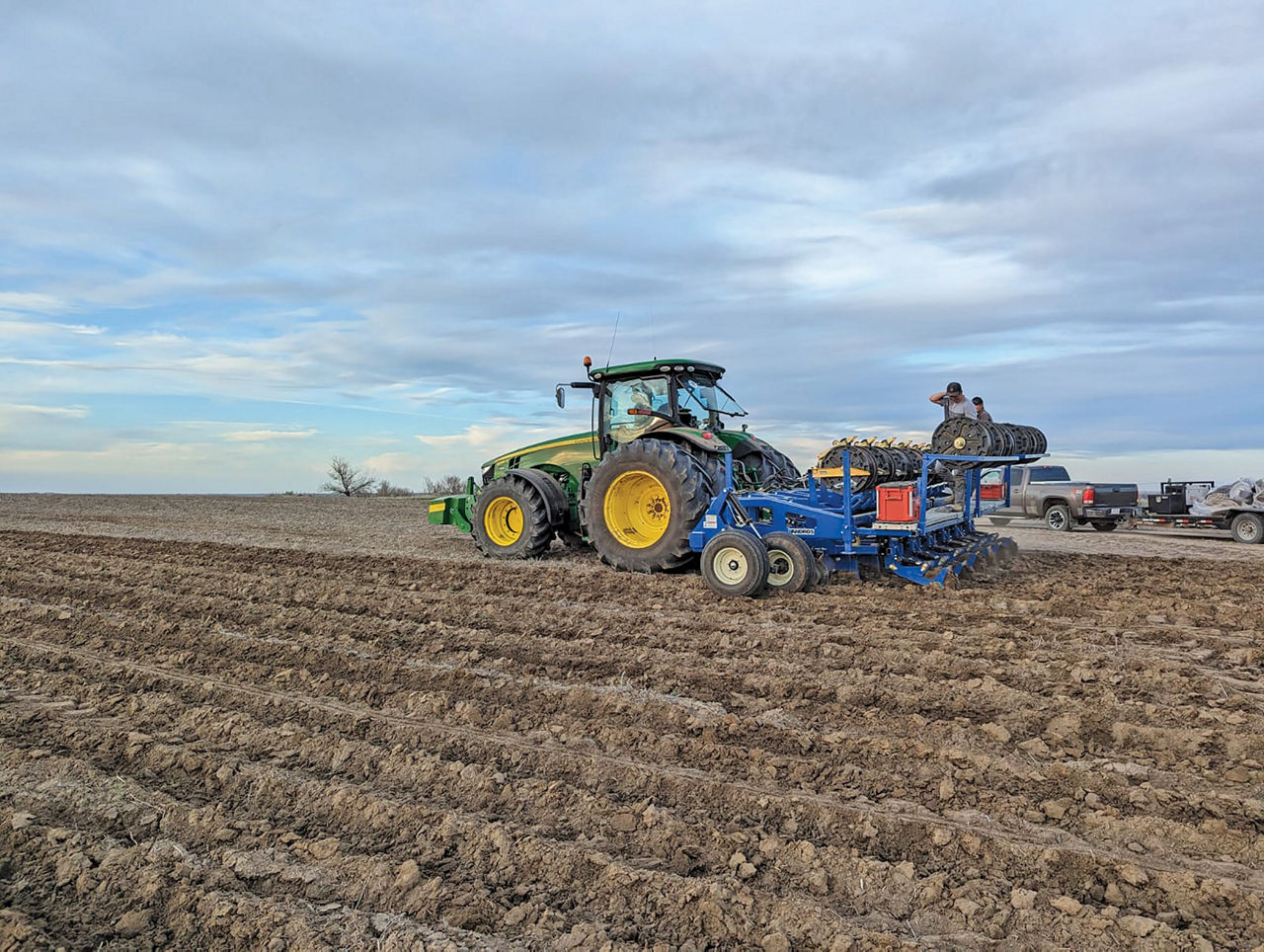 A green tractor pulls a blue agricultural implement across a freshly plowed field under a cloudy sky.