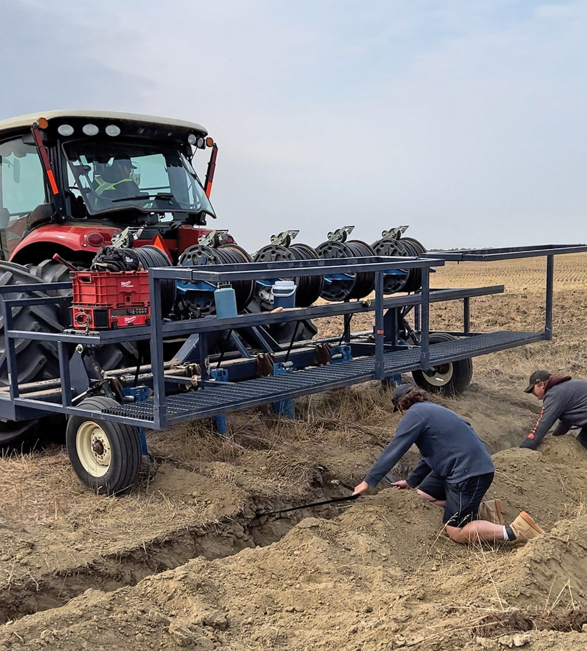 A red tractor with a metal platform carrying large spools of tubing and equipment is parked in a field. Two people are kneeling nearby, working in a trench in the soil. The field appears recently plowed and stretches into the distance under a cloudy sky. 