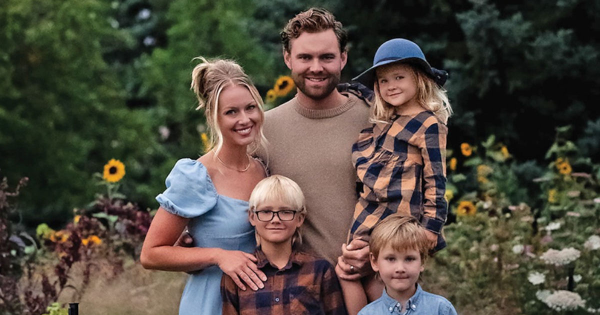 A family of five stands close together in a lush garden surrounded by colorful flowers. The parents stand behind their three young children, and they are all smiling at the camera.  