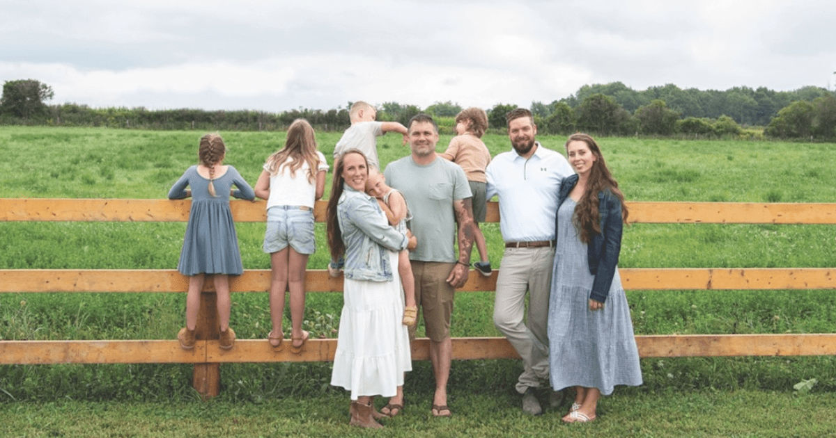Two couples pose for a photo in front of a wooden fence. Several children climb on the fence behind them. 