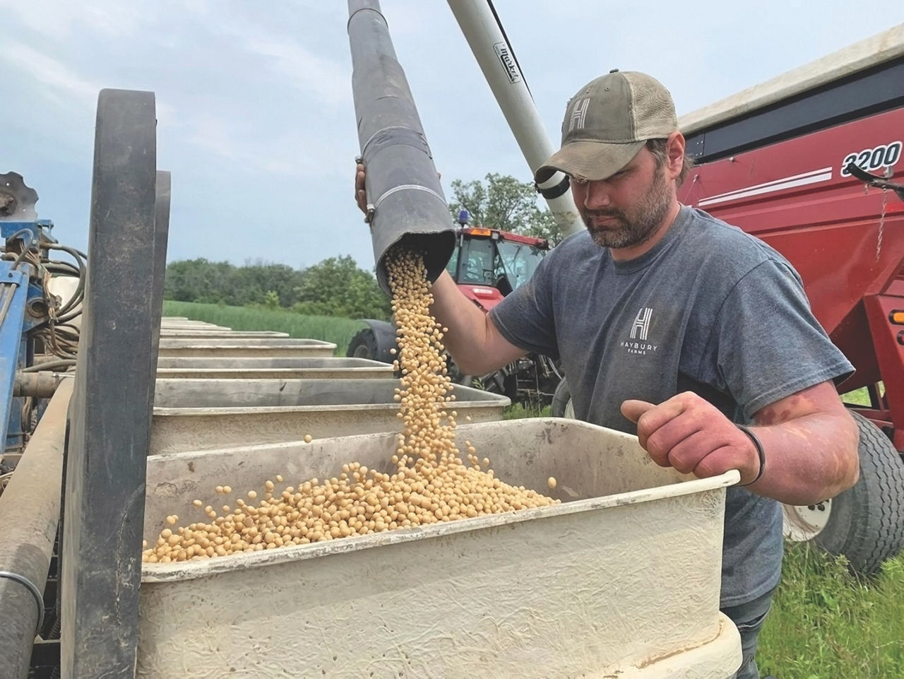 A man stands in front of a large bin holding a nozzle coming from a truck. Soybeans are coming out of the nozzle into the bin. 