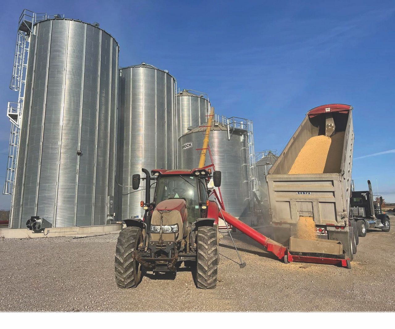 Large steel silos stand tall while a tractor siphons corn from a dump truck into one of the smaller silos.