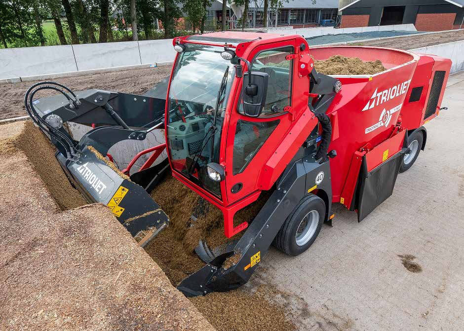 Trioliet feed mixer machine in red. Cattle feed is in the back of the mixer and is being pushed into stacks at the front of the machine, operated by one person in the cab. Barns and trees in background.