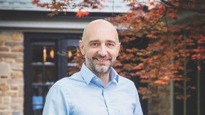 A man standing in front of a brick home, smiling with his hands in his pockets. He is wearing a blue button up shirt. 