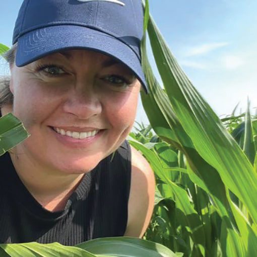 Close up image of Ashley Smith wearing a dark sleeveless shirt and blue cap standing among tall green corn plants under a clear sky. 