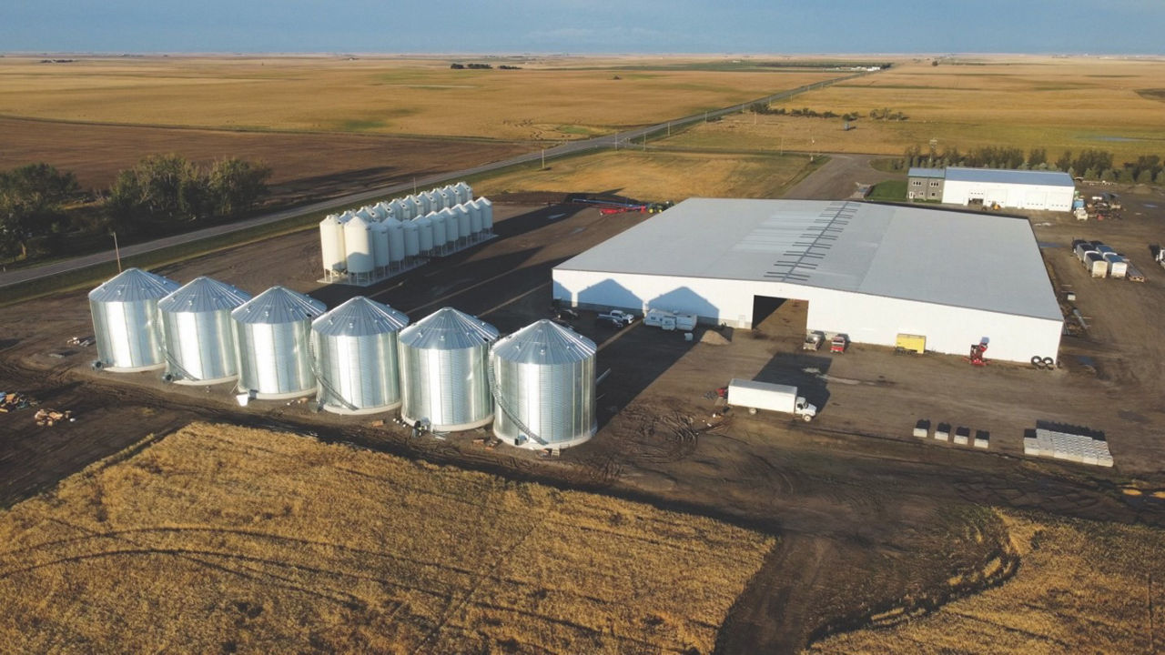 An aerial view of six large silver silo bins on a farm. Nearby is a large white warehouse and some smaller white silos. 