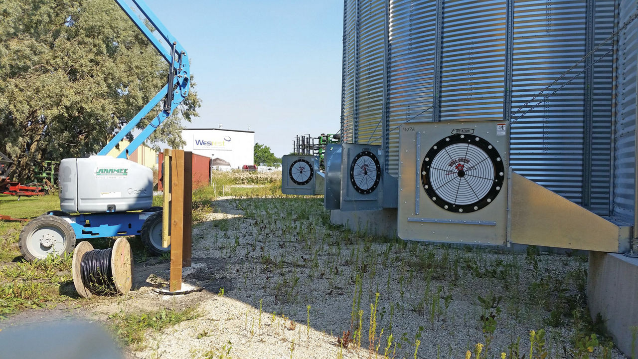 Grain silo with three ventilation fans, a aerial lift nearby, and a spool of cable on gravel ground in an industrial setting.
