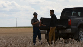 Two farmers standing behind a black pickup truck in stubble.  