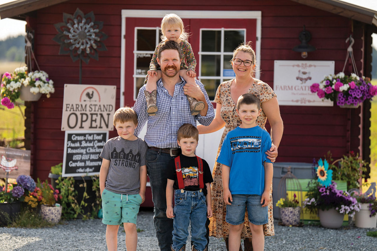 Cuthbert family standing in front of a rustic red farm shop with flowerpots and signs, promoting a local farm business and fresh produce.