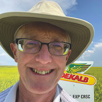 A smiling man, Dave Burns of Canola Bayer, is pictured in a sunhat standing in front of a yellow crop of canola. The DEKALB logo is shown behind him.