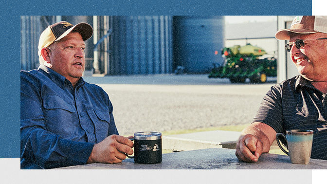 A person with a hat sitting at a table holding a mug in front of a combine and storage. 