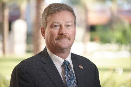John Schmeiser smiles at the camera as he poses for a portrait. He has a moustache and is wearing a suit and tie. 