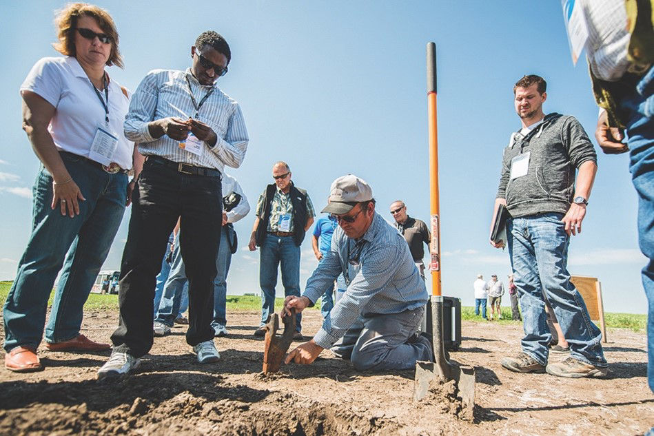 A group of agronomists standing in a field on a blue-sky day, observing a man crouched down pointing to the soil.