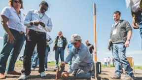 A group of agronomists standing in a field on a blue-sky day, observing a man crouched down pointing to the soil. 