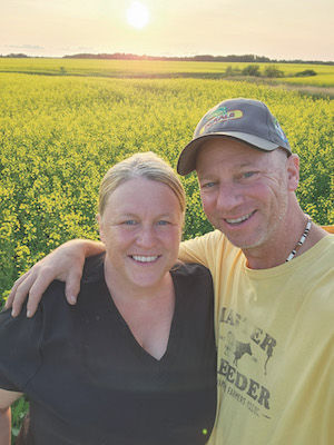 A smiling man and woman stand in front of a healthy green field with golden sunlight setting on the crop.