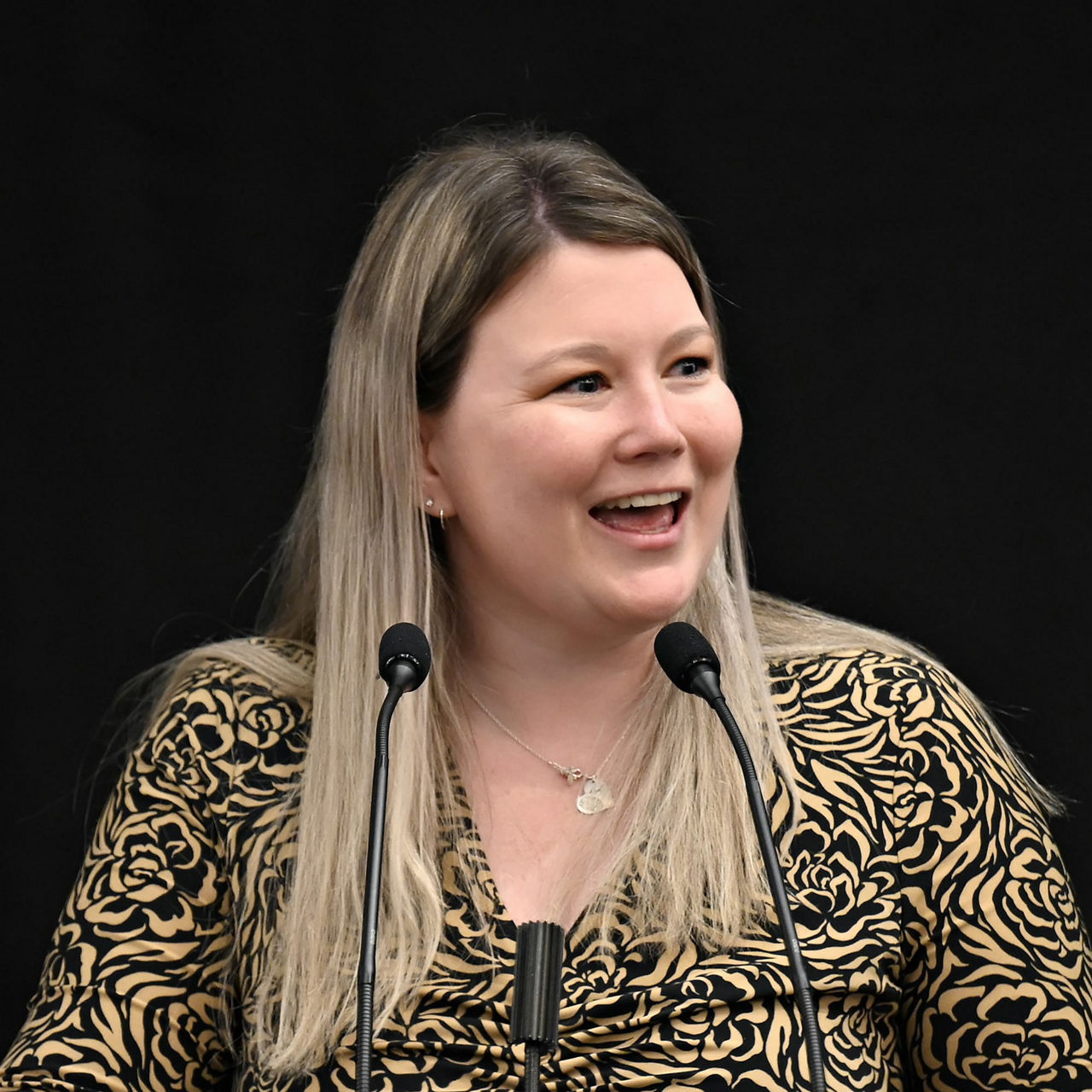 Heather Watson speaking at a podium with two microphones against a black background. 