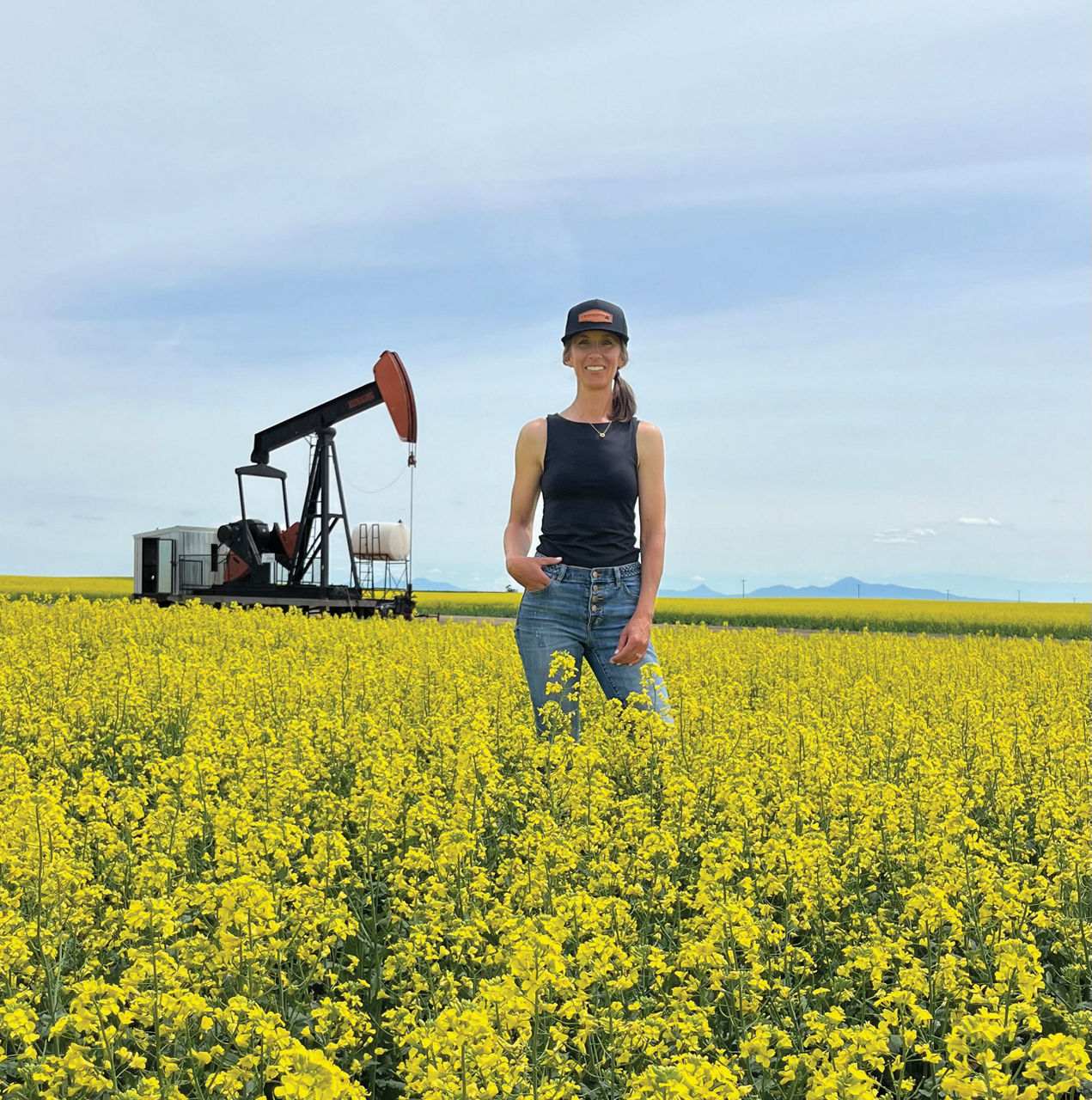 Jenny Seward standing in a blooming yellow canola field with an oil pumpjack in the background under a cloudy sky. 