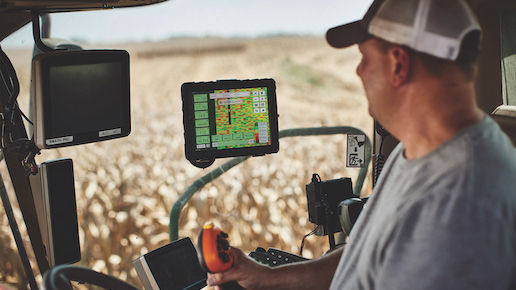 A farmer sits inside a tractor cabin while looking at a tablet screen. The tablet is showing the FieldView software.