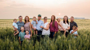 A large Caucasian family of seven adults and six children stand in a sprawling field of wheat, with silos along the distant horizon. 