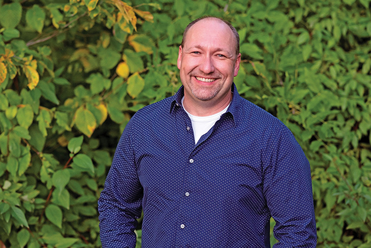 Headshot of a man in a blue button up shirt in front of a leafy plant background. 