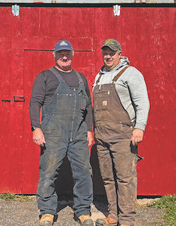 Two men in farming coveralls standing in front of a red barn