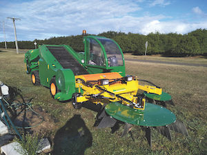 A large green tractor sits on green grass, and the tracker a macadamia harvester with a large front pronged section.