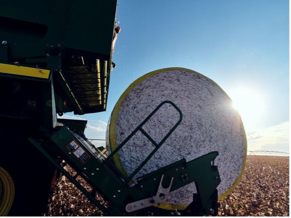Close-up of a cotton bale being harvested by a green harvester in a field, with the sun setting in the background. 