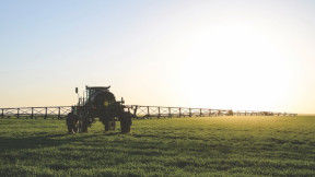 A large tractor with a wide sprayer drives along a flat field that sprawls into the horizon. The sun is setting in the distance.