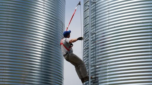 Farmer climbs a ladder up the side of a grain bin, clipped onto a safety line with a harness.