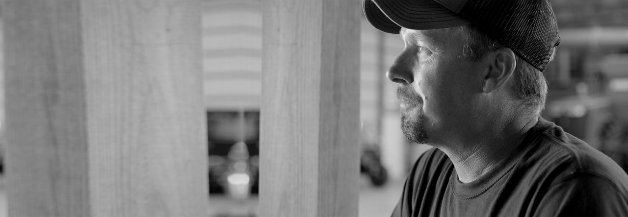 Black and white side profile of a farmer in a cap, looking out from inside a building. 